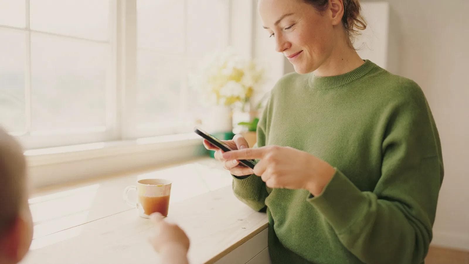Kobieta w zielonym swetrze pisze wiadomość w telefonie przy jasnym kuchennym blacie, obok kubek herbaty i rozmyta rączka dziecka. Spokojna, jakby właśnie umówiła wizytę