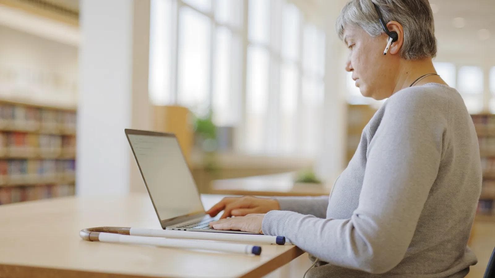 Niewidoma kobieta w szarym swetrze ze słuchawką w uchu pewnie pisze na klawiaturze laptopa w jasnej czytelni biblioteki. Obok na drewnianym stole leży złożona biała laska
