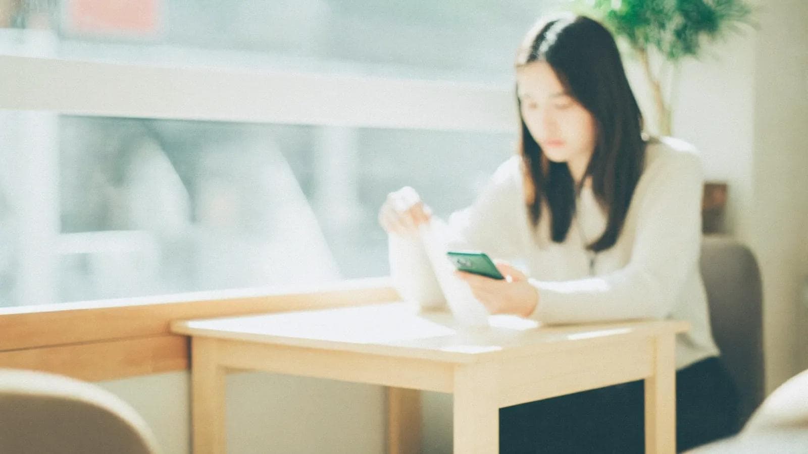 A woman in a light sweater at a pale wood table in a sunny café quickly finishes something on her phone